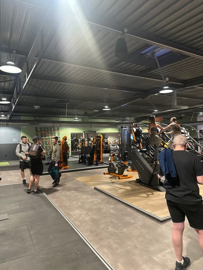 This image captures the interior of a gym in Annœullin, France, featuring several individuals engaged in fitness activities. The setting showcases a modern, industrial aesthetic with a high, corrugated metal ceiling supported by exposed beams, illuminated by numerous circular overhead lights and some directional spotlights. Large black speakers are mounted on the walls near the ceiling.

In the foreground and mid-ground, several people are using cardio equipment. On the right, two individuals are seen from the back, actively using stair climber machines. One person wears a black t-shirt and shorts, with a blue towel draped over their shoulder, next to a machine clearly marked with the "MATRIX" brand logo. Another person, further back, wears a patterned top and shorts while operating a similar machine. In the deeper background, more fitness equipment with orange accents, including exercise bikes, is visible.

The floor of the gym features a blend of light wooden-look panels and darker grey tiled sections. Towards the left side of the frame, a group of three men is standing, one of whom is smiling towards the camera, wearing a light grey hoodie and a backpack. Another person in a dark jacket stands further back. The overall scene depicts a busy, functional gym environment during what appears to be a regular operational period, though the specific time of day is not discernible from the indoor lighting.