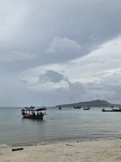 The image depicts a cloudy day scene at Koh Toch Village, Cambodia. The foreground features a sandy beach with a piece of driftwood and some scattered debris, including a plastic bottle. The sea is calm with gentle waves lapping at the shore. Several small fishing boats are anchored on the water, some closer to the shore and others further out. In the distance, a forested island or landmass is visible under the overcast sky. The overall atmosphere is serene, with the cloudy weather creating a diffused light.