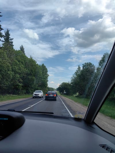 This is a vertical, eye-level, daytime, full-shot photograph taken from the interior of a vehicle traveling on a paved road. The road runs through a rural landscape with dense green trees lining both sides. The sky is partly cloudy with scattered white and grey clouds against a light blue backdrop.
In the foreground, the dashboard of the vehicle is visible, along with a portion of the windshield frame on the right. On the road ahead, several cars are visible. A white SUV is in the left lane, and a dark-colored sedan is in the right lane, followed by another dark-colored vehicle. The cars are traveling in the same direction as the photographer.
The scenery suggests a journey through a forested or heavily treed area. There is a yellow diamond-shaped road sign with black symbols on the right side of the road. The environment appears to be the territory of Ilyanskiy selskiy Sovet, Belarus, given the presence of a sign with Cyrillic text that reads "Ільянскі сельскі Савет" (Ilyanskiy selskiy Sovet) and "Вязынка" (Viazynka), indicating the geographical location.