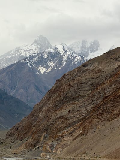The image shows a dramatic mountain landscape under a cloudy sky. In the foreground, a steep, rocky mountainside dominates the right side of the frame, with textured brown and tan rock formations. To the left and in the background, successive layers of mountains recede into the distance. The further mountains are darker and have sharp, jagged peaks, some of which are capped with snow. The sky is overcast with gray clouds. The scene appears to be a remote, high-altitude environment. There are no people or man-made objects clearly visible in the image, except for a faint hint of a road or path at the very bottom of the frame. The absence of specific landmarks or distinguishing features makes it impossible to pinpoint the location within Stongde, India, or any other city. The image focuses solely on the natural geological features of a mountainous region.