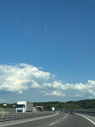 This image shows a highway scene with a clear blue sky and white clouds. The highway appears to be a multi-lane road with a guardrail separating it from the adjacent landscape, which is filled with lush green trees. Several vehicles are visible on the road, including a white truck with a trailer, a red and black truck, and a few cars. The vehicles are traveling in the same direction. The perspective suggests the photo was taken from another moving vehicle. The weather appears to be sunny and pleasant, suggesting it's daytime. There are no visible people, but the presence of vehicles indicates human activity. The location is identified as Baliovtsi, Bulgaria, which would imply a European setting. There is some text visible on the white truck's trailer, but it is too blurry to read. The overall impression is a typical highway scene on a clear day.