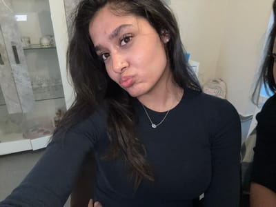 A young woman with dark hair and brown eyes is taking a selfie in a room. She is wearing a long-sleeved black shirt and a silver necklace with a blue pendant. Her lips are pursed, and she has a small pearl earring in her left ear. In the background, there is a display cabinet with decorative items and glassware. To the right, another person is partially visible, wearing a black top. The room appears to be a domestic setting. The image was taken in Maldegem, Belgium.