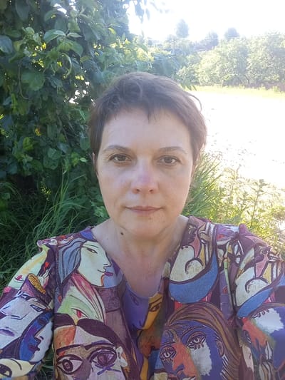 A selfie of a woman with short, dark brown hair and light skin is taken outdoors. She is wearing a colorful shirt with a cubist-style print featuring various faces. The woman's face is looking directly at the camera. Behind her, there is lush green foliage on the left and a bright, sunlit field with trees in the background on the right. The environment suggests a rural or suburban setting, possibly a garden or park, with hints of houses and fences in the distance, indicative of the village of Retun, Russia.
