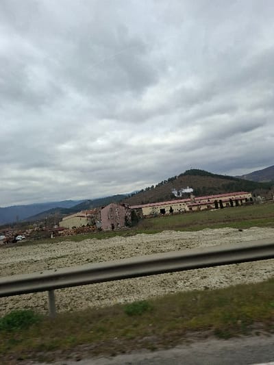 The image depicts a rural-industrial landscape in Cherniche, Bulgaria, under an overcast sky. In the immediate foreground, a metallic guardrail runs horizontally, partially obscuring a strip of green grass and a disturbed, light-colored field beyond it. The midground features a cluster of buildings: to the left, a multi-story structure with faded pink plaster; to its right, a longer, lower industrial complex characterized by reddish-brown roofs and lighter walls, with at least one chimney actively emitting a plume of white smoke. Adjacent to these buildings and to the left, large stacks of what appear to be lumber or processed materials are visible, along with several white vehicles. The background consists of rolling, tree-covered hills and mountains extending into the hazy distance under a uniformly gray, cloudy sky. A faint contrail is visible in the upper central sky. No discernible individuals are present in the scene.