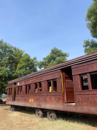A dark brown railway carriage with the yellow letters "WR" on its side sits on wheels in a grassy area. Trees and blue sky are visible in the background. The carriage is located in Anajpur, India.