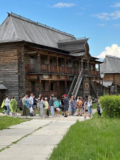 The image captures a scene at the Vzglyadnevo estate, Russia, featuring a large wooden structure beside a paved pathway. A group of approximately 20 people stand in a line along the path, most facing towards the building. The structure appears to be a multi-story wooden building with intricate wooden detailing and multiple levels connected by exterior wooden stairs. The building is weathered and the wood is a gray-brown color. A patch of green grass is visible to the right of the pathway, adjacent to the group of people. The sky above is blue with some white clouds visible.