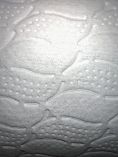 The image shows a close-up of a textured white surface, possibly wallpaper or fabric, with an embossed pattern of curved lines and small circular indentations. The lighting creates shadows that highlight the three-dimensional nature of the design, suggesting a raised pattern against a slightly textured background. There are no people, objects, or clear environmental context to indicate a specific location within Letterkenny, Ireland. The image focuses solely on the tactile and visual qualities of the material's surface.
