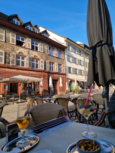 The image captures an outdoor setting, possibly a café or restaurant in Rheinfelden, Switzerland. The camera angle is low, directed upwards towards a building with distinctive architectural details. The building is several stories high and features painted decorative elements, windows with shutters, and visible signage in German. A man is leaning out of one of the windows. In the foreground, there is a table setting with drinks (one with a pink straw), a dish of what appears to be fries, and a menu or brochure. The seating arrangements consist of chairs around the table. Above the table, there is a partial view of a sun umbrella. The sky is a clear, bright blue. The scene suggests a leisurely outdoor experience with a view of the building's architecture. The printed text on the building reads: "bis 1884 Braustätte des Rheinfelder Urbräu Dormals Dietschy's Bierhaus."