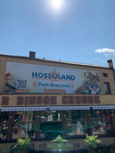 The image shows the facade of a building with a large banner advertising "HOSSOLAND Park Rozrywki". Below the banner, the name "ROBINSON CRUSOE" is prominently displayed in large, brown, three-dimensional lettering. The building has a brown exterior with large windows framed by wooden beams. A blue and white striped awning stretches across the top of the windows. In the windows, reflections of trees and people are visible, as well as posters and what appears to be a Red Bull neon sign. In front of the building, there are tables and chairs, and potted palm plants. The sky above is clear blue with a few scattered clouds, and the sun is bright, causing some lens flare. The building is located in Rewal, Poland, as indicated by the banner.