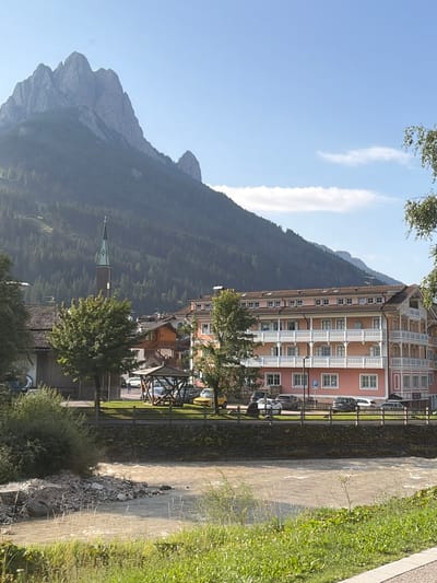 The image depicts a scenic view of Pozza di Fassa, Italy, with a prominent mountain range in the background. A tall, slender church steeple with a green spire rises above the rooftops of a charming town. A multi-story pink building with white balconies and numerous windows dominates the middle ground, suggesting a hotel or residential complex. In the foreground, a fast-flowing river, the Avisio, meanders through the landscape, its waters a muddy brown color. The riverbanks are lined with green grass and some rocky debris. Several cars are parked along the street in front of the pink building, and a wooden gazebo-like structure stands near the river. Trees are scattered throughout the scene, adding to the natural beauty of the location. The sky is a clear blue, with a few wispy clouds.