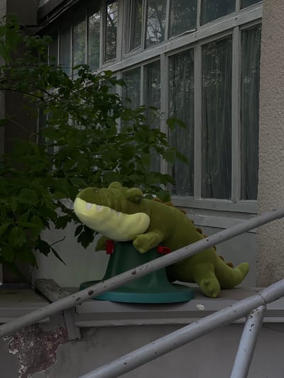 A stuffed green alligator toy is mounted on a green, trumpet-shaped object attached to a weathered wall with peeling paint, possibly in Bobrov, Russia. A leafy green plant is visible in the foreground. A metal staircase and a windowed structure are also present in the background.