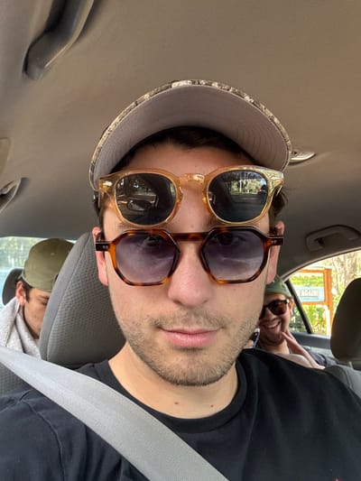 The scene depicts the interior of a vehicle during daylight in Micanopy, United States, occupied by at least three male individuals. In the foreground, a male adult is positioned as if taking a selfie, wearing a grey camouflage-patterned baseball cap, a black t-shirt, and a grey seatbelt across his chest. He is wearing a lower pair of tortoiseshell-patterned hexagonal sunglasses with dark tinted lenses, and an upper pair of amber-framed round sunglasses perched atop his head, reflecting a tree-lined road and distant vehicles. His face exhibits stubble, and his expression is neutral with closed lips.

Behind the main subject, on the left, a second male adult is partially visible, wearing an olive-green baseball cap and appearing to be resting or leaning forward, with a light-colored cloth draped over his shoulder. On the right, in the rear passenger seat, a third male adult is visible, wearing a green baseball cap and sunglasses, smiling broadly with his right hand raised towards his face. Through the rear passenger window, a brown commercial sign with white lettering is discernible, prominently featuring the word "AGAIN!". The vehicle's interior headliner is a light beige or grey.