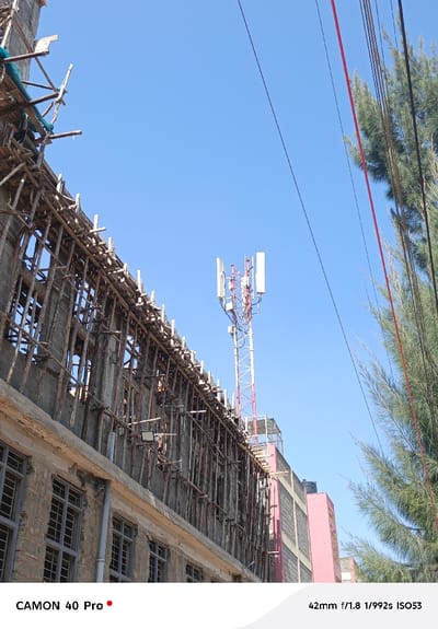 A daylight, low-angle urban scene in Oloosirkon/Sholinke ward, Kenya, depicts a multi-story building under construction or renovation on the left. This structure features exposed concrete block walls on lower floors with grid-patterned windows and extensive wooden scaffolding on upper levels, partially covered by green netting. An LED floodlight is affixed to the building's wall. To the right, a red and white lattice telecommunications tower, equipped with multiple cellular antennae, rises above surrounding structures. Further right, a dense cluster of tall, slender green trees partially obscures another building with pink and grey facades. Multiple black and red utility lines traverse diagonally across the upper right portion of the frame. The sky is clear blue. No individuals are visible.