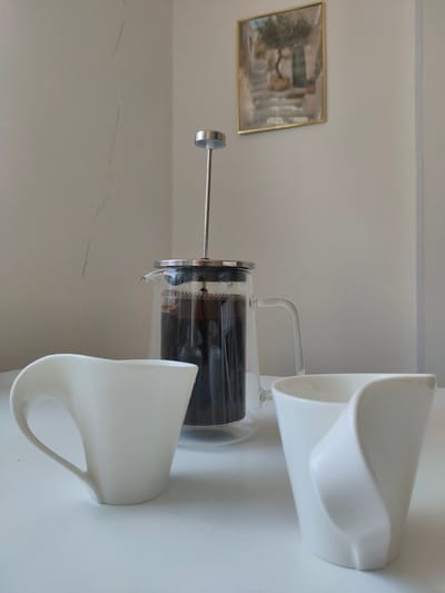 The image shows a table setting with a French press coffee maker and two white ceramic mugs. The French press is made of glass and metal, and it contains dark coffee. The mugs have a modern, sculptural design. In the background, a framed picture of a Mediterranean-style scene with an olive tree hangs on a light-colored wall. The wall has a faint crack running vertically down it. The scene appears to be indoors, possibly in a home or a cafe. There are no people visible in the image. The overall impression is one of a peaceful, quiet moment, perhaps a break for coffee. There is no information that would place this scene in Zrenjanin, Serbia.