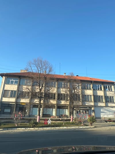 The image captures a static, exterior view of a large, multi-story building in Resilovo, Bulgaria, under a clear blue sky. The structure presents a light-colored, likely stucco or concrete facade with numerous rectangular windows across three visible floors, topped by a red terracotta tile roof. Several bare deciduous trees stand in front of the building, partially obscuring the facade. Utility lines traverse the upper portion of the scene.
A central entrance on the ground floor features a partially legible sign, with "КМЕТСТВО" visible, indicating a mayoral office or municipality building. The ground level also contains several shop-front style windows, some appearing to be covered or vacant.
In the landscaped green space directly in front of the building, various red and white decorations are prominently displayed. These include two large, stylized figures, one primarily red and one primarily white, positioned on the grass. Numerous smaller red and white ribbons and ornaments are tied to the branches of the smaller trees and shrubs, consistent with traditional Bulgarian Martenitsi. Some tree trunks are wrapped with red and white striped fabric.
The immediate foreground shows a section of a dark asphalt road, with a partial reflection of the observer's vehicle on its surface. No individuals or specific actions are discernible within the frame. The lighting suggests a bright, sunny day.