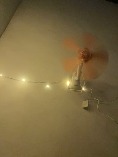 The image depicts a ceiling fan with attached fairy lights. The fan, primarily pink in color, is in motion, evidenced by the blurred appearance of its blades. A white electrical component, possibly a light or a sensor, is affixed to the fan's motor housing. The text "SOES" is visible on this component. A string of small, warm-toned LED fairy lights is draped around the fan and extends downwards, creating a festive or decorative atmosphere. The lights are illuminated. A white wire connects to the electrical component on the fan, and another wire runs towards a small white square object mounted on the ceiling, which could be a light switch or a junction box. The overall scene is dimly lit, with the primary illumination coming from the fairy lights and possibly a light source not directly visible in the frame. The ceiling appears to be painted a neutral, light color with some visible texture and minor cracks. The image is taken from a low angle, looking up at the ceiling. The setting appears to be indoors, likely a room. The time of day is suggested to be evening or night due to the artificial lighting and the ambiance created by the fairy lights.