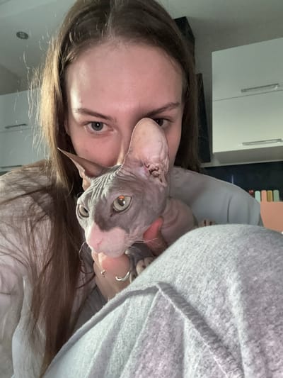 A close-up, low-angle, indoor selfie shows a woman holding a hairless Sphynx cat. The woman has long brown hair and is wearing a white shirt and grey sweatpants. She is looking at the camera with a neutral expression, and her left hand, adorned with two rings, is gently holding the cat's face. The cat, predominantly grey with pinkish undertones on its nose and ears, has large, light green eyes and prominent wrinkles. The background is blurred, showing kitchen cabinets and a wall with some colorful objects. The scene suggests a domestic setting, possibly in Novaya Ivanovka, Russia.