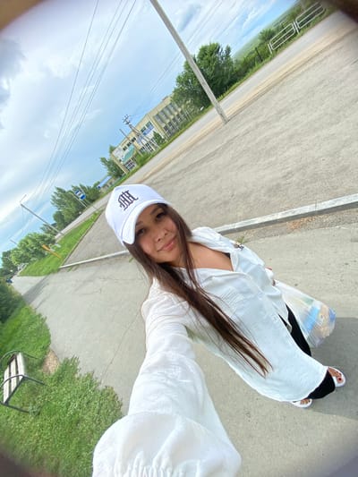 A young woman with long, dark hair is taking a selfie outdoors. She is wearing a white baseball cap with a dark embroidered logo, a white button-down shirt, and black pants with white sandals. She is holding a clear plastic bag filled with items. The background shows a paved road, a sidewalk, a grassy area with a bench, trees, a building with many windows, and a utility pole with wires. The sky is partly cloudy. The location is the city of Akyar, Russia.