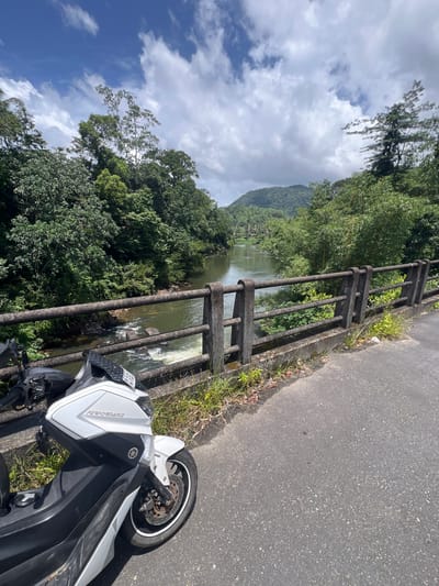 A white and black scooter is parked on the side of an asphalt road that crosses a bridge over a calm river. The bridge has a concrete railing with horizontal metal bars. Lush green trees and vegetation line the riverbanks and extend up the hills in the background. The sky is partly cloudy with patches of blue. The scooter is angled towards the river, with its front wheel visible. The scene appears to be a scenic spot in Panangala Dhahe kade, Sri Lanka.