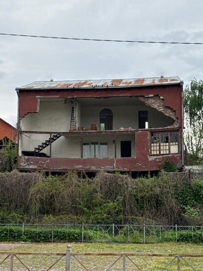 The image shows the ruined shell of a three-story building in Assinovskaya, Russia. The front wall has partially collapsed, exposing the interior floors and staircases. The remaining facade is painted in a weathered red hue. Windows are either broken or missing. Overgrown vegetation encroaches on the building's right side. A section of a decorative metal fence is visible in the foreground. The sky is overcast.