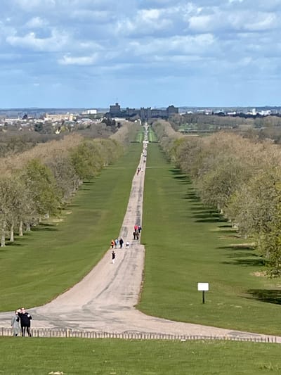 An elevated, long-distance view captures a wide, expansive park landscape under a partly cloudy sky. A long, straight, light-colored paved path extends from the foreground into the far distance, flanked on both sides by extensive green grass and parallel rows of deciduous trees with minimal foliage. At the distant end of the path, the distinct silhouette of Windsor Castle is visible, a large stone complex with multiple towers, surrounded by a townscape.
In the foreground, on the left, a group of three individuals stands on a grassy area behind a low, light-colored wooden post-and-rail barrier. The central figure wears a dark jacket, one to their left wears a light grey coat, and the third is partially obscured. To the right of the path in the foreground, a plain white rectangular sign on a dark pole is planted in the grass.
Along the main paved path, numerous individuals and small groups are engaged in walking. Notable groups include 3-4 people in the lower-midground, one person wearing a red jacket and another an orange top, with at least one dog accompanying them. Further along the path, other figures appear as small dark shapes. Shadows from the trees are cast to the right side of the path, indicating a light source from the left. This scene is located in the city of The Village, United Kingdom.