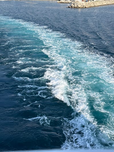 The image shows the turbulent, foamy wake of a vessel cutting through deep blue water, departing from the port of Kamariotissa, Greece. The wake, a mixture of white froth and lighter blue water, diagonally bisects the frame from the lower right to the upper left. In the upper background, the man-made rock breakwater of the Kamariotissa port is visible, constructed from light-colored boulders. Behind this breakwater, several small white boats or yachts are docked, and indistinct human figures are discernible on the distant shoreline within the harbor area. The overall environment is open sea and a coastal port under daylight conditions. No specific individuals or direct interactions are clearly identifiable on the vessel creating the wake.