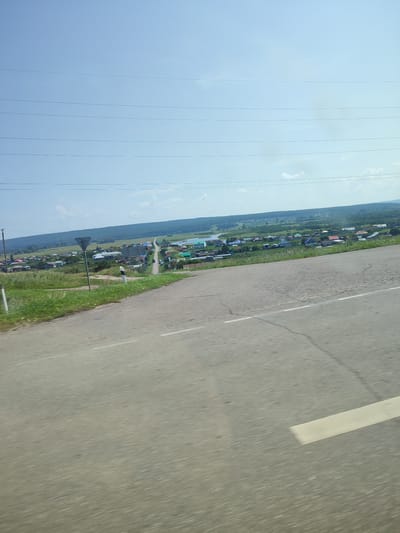 This is a medium shot, eye-level, horizontal, outdoor, daytime, natural-light, still photograph captured from a moving vehicle. The image depicts a rural landscape with a paved road in the foreground. The road is grey with a white line on the right side. Beyond the road, there is a green grassy slope leading down to a village. The village consists of numerous houses with colorful roofs, nestled among trees. A few power lines are visible in the sky. The sky is a clear, light blue. The scene suggests a tranquil, rural setting in the village of Lemez-Tamak, Russia.