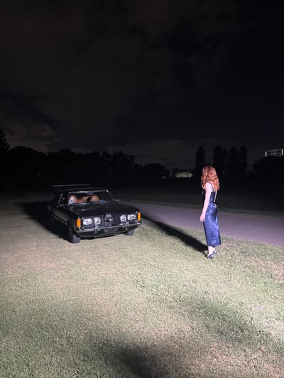 A dark-haired woman in a blue jumpsuit stands next to a black car parked on a grassy field at night. The grass is illuminated by a light source, casting a long shadow from the woman. The sky is dark and cloudy. Trees are visible in the background, and a structure that appears to be power lines can be seen in the distance.