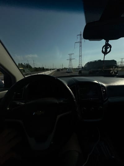 A first-person perspective captures a view from inside a car on a highway. The steering wheel and dashboard are in the foreground, dark and silhouetted. Through the windshield, tall electrical transmission towers line the side of the road, with power lines stretching across the clear blue sky. Other vehicles are visible on the highway in the distance. The scene appears to be set in a suburban or rural area, possibly along a major route. The image does not contain any specific identifying features of the city of Istiqlol, Uzbekistan.