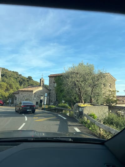 Captured from inside a vehicle traveling along a narrow road, the image reveals a street view of the medieval village of Èze, France. A dark-colored car is visible on the right-hand side of the road, with a red vehicle partially visible further down the road. The architecture features stone buildings, one with a small church and cross on top. Trees with dense foliage and an olive tree are present along the road. Yellow lines on the asphalt indicate parking restrictions. The sky is blue with light clouds. The image is taken from the perspective of the driver or passenger of a car, with parts of the interior of the vehicle visible on the sides of the frame.
