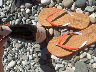 In Amzara, Abkhazia, a bottle of wine with a label reading "Абхазия" sits on a beach of grey and white pebbles. Next to the bottle are two pairs of flip-flops with orange straps and wooden soles, one pair with a white sole and the other with a brown sole. The scene is lit by sunlight, casting shadows from the pebbles.
