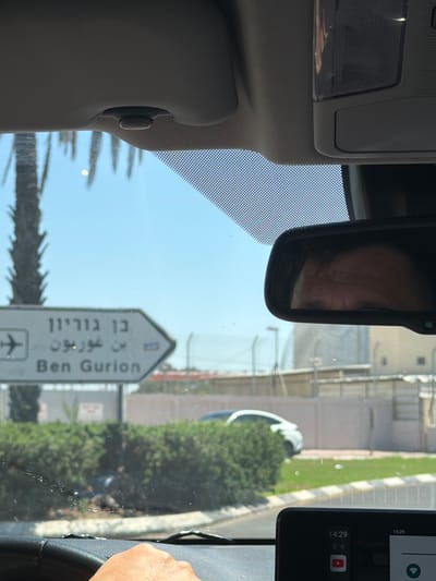 The image is taken from inside a car, looking forward through the windshield. The driver's left hand is visible on the steering wheel. A signpost with an airplane icon and text in Hebrew and English points to the right, indicating "Ben Gurion." The background shows a clear blue sky, some palm trees, a green hedge, and a building with a fence. The interior of the car is visible, including the rearview mirror reflecting a person's face, the sun visor, and the dashboard with a screen displaying the time "14:29" and icons for YouTube and other applications. The environment appears to be sunny and outdoors. The signpost confirms the location is in Kfar Truman, Israel, as it directs to Ben Gurion, which is the major international airport serving Tel Aviv and is located near Kfar Truman.