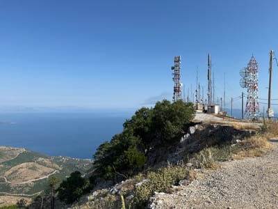 This media file captures a wide-angle view from a mountaintop in the Kassopaia Municipal Unit, Greece. The scene is dominated by a clear, bright blue sky. In the foreground, a rough, gravelly path leads up a rocky slope covered in sparse, dry vegetation and scattered green bushes and trees.

The midground of the image is characterized by a cluster of telecommunications towers, varying in height and design. Some are lattice towers, painted red and white, while others are simpler metal structures. They are surrounded by some buildings and more vegetation, including a dense cluster of trees on the left side of the frame. The towers suggest a functional, perhaps industrial, use of this elevated location.

In the background, a vast expanse of deep blue sea stretches towards a distant coastline, which appears hilly and dotted with sparse settlements. The water is calm, with a faint shimmer indicating the bright sunlight. The distant landforms are hazy, suggesting a considerable distance.

The time of day appears to be midday, with the sun high in the sky, casting sharp shadows. The weather is clear and sunny, ideal for the panoramic view offered. The location cues like the Mediterranean Sea, the vegetation, and the mountainous terrain are consistent with a Greek island or coastal mountain region. There are no people visible in the image, and no discernible text is present. The overall impression is one of a remote, elevated communication hub overlooking a scenic coastal landscape on a clear, sunny day.