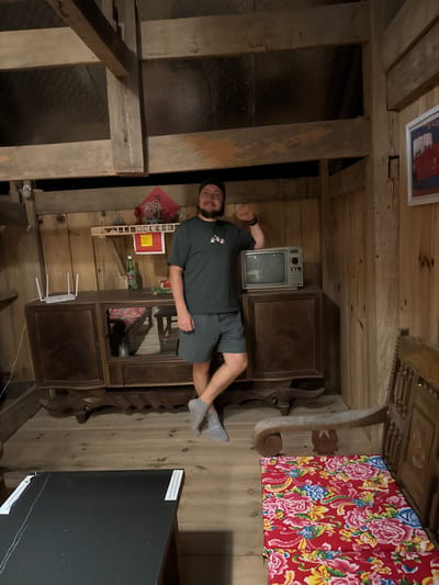 A horizontal shot reveals a man lying on an ornate, wooden cabinet in a room with wooden walls and ceiling. The location is the Hoàng Su Phì Commune in Vietnam.

The man is wearing a dark green t-shirt and grey shorts. He is lying on his side, facing the camera, with one arm outstretched.

The wooden cabinet is intricately carved and sits against a wooden wall. On top of the cabinet are a vintage television, a red flowered quilt, and a wooden sculpture. There are items on shelves in the cabinet including a router.

The room is lit with a warm, dim light, creating a rustic atmosphere. The ceiling is made of wooden beams, and the walls are also clad in wood. In the background, an opening in the wall is visible.