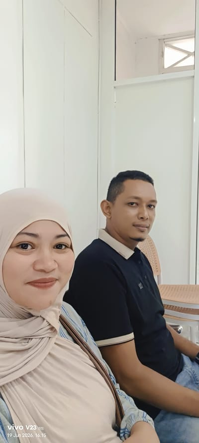 The image shows two people, a woman and a man, seated indoors. The woman is in the foreground on the left, wearing a beige hijab and a light-colored top with a blue pattern. She is looking towards the camera with a slight smile. The man is seated behind her and to the right, wearing a black polo shirt with a light-colored collar and trim. He is looking slightly to his left, away from the camera. The background appears to be a brightly lit room with white walls and what seems to be vertical paneling. There are chairs visible in the background, one of which the man is sitting on. The overall impression is of a candid photograph taken in a public or waiting area. The timestamp on the image indicates "19 Jun 2026, 16.15", and "vivo V23 5G" branding is also present.