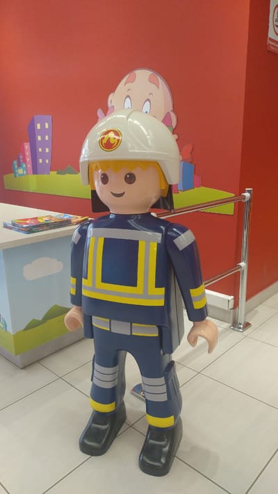 A large, plastic, toy-like firefighter figure stands in an indoor space with a red wall and tiled floor. The figure has a blue uniform with reflective yellow and gray stripes on the chest, sleeves, and legs. It wears a white helmet with a red and yellow emblem. Behind the figure, a colorful mural depicts abstract buildings and green hills. A counter with books is to the left, and a chrome railing with a red bar is to the right. The overall scene appears to be a play area or promotional display. The specific location in Pylaia Municipal Unit, Greece is not determinable from the image.