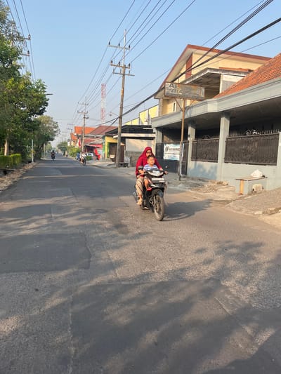 The image depicts a street scene in Sugih Waras, Indonesia, bathed in the warm light of a late afternoon sun. The asphalt road is well-maintained, with some patches indicating previous repairs. On the right side of the road, a gray building with a dark, ornate metal fence stands. A small wooden bench is visible outside this building, with a white plastic bag placed on it. Further down the street, several buildings with terracotta-tiled roofs line both sides, suggesting a residential or commercial area. Electrical poles and wires crisscross the sky, a common sight in Indonesian urban landscapes.
The primary focus of the image is a woman and a child riding a motorcycle, traveling away from the viewer. The woman, wearing a red headscarf and a patterned skirt, is driving. The child, dressed in a blue and white striped shirt, is seated in front of her. Both appear relaxed. In the distance, other motorcycles and pedestrians can be seen, indicating a moderate level of activity on the street. Trees with lush green foliage line the left side of the road, casting long shadows across the asphalt. The sky is a clear, pale blue. The overall impression is of a typical day in this Indonesian town, with everyday life unfolding on its streets.