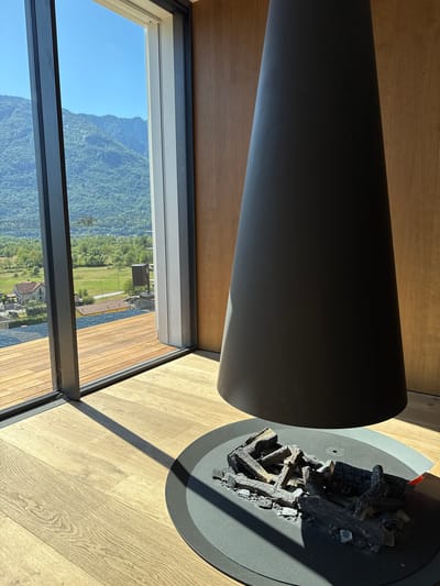 This media file captures an indoor scene with a large, cylindrical, dark gray object dominating the foreground, likely a modern fireplace or heater, positioned on a light-colored wooden floor. To the right of this object, a circular, dark gray base holds what appears to be artificial or used firewood logs, suggesting it is a fireplace. Sunlight streams in from the right, casting a strong shadow across the floor and the fireplace.
Through a large window that spans the lower portion of the image, a lush green landscape is visible. Rolling hills and mountains covered in dense vegetation are prominent under a clear blue sky. Hints of man-made structures, possibly houses or buildings, are scattered within the landscape. The light wood flooring and the expansive window with a view of the natural scenery suggest a modern dwelling in a scenic, possibly mountainous, location.
The scene is set during the daytime, with bright sunlight indicating clear weather. The overall impression is one of modern interior design harmoniously integrated with a natural outdoor environment. No people are visible, and there is no discernible text. The setting suggests a tranquil and serene atmosphere.