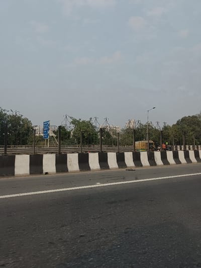 This image presents an outdoor scene captured from an elevated perspective, likely from a moving vehicle, on an overcast or hazy day in Ghaziabad, India. The foreground is dominated by a multi-lane asphalt road, featuring a continuous white lane marker. To the left of the road is a concrete barrier, distinctly painted with alternating vertical black and white stripes.
Beyond this barrier, an ornate black metal fence, adorned with decorative scrollwork along its top, extends horizontally across the frame. Behind the fence, a dense line of green trees and shrubs partially obscures a lower-level area, possibly another road or service lane. Through the foliage and fence on the left side, a blue rectangular road sign with white Hindi text is visible, although largely unreadable. It appears to be a directional sign, possibly indicating "Ghaziabad" and a distance such as "2km" or "20km."
Towards the right, a street light pole with a modern light fixture rises above the fence line. Directly beneath it, on the lower level, a yellow auto-rickshaw is clearly visible, either parked or stopped. Around the auto-rickshaw, several indistinct figures, likely people, and possibly other small vehicles like motorcycles, are gathered. In the distant background, through the hazy atmosphere and gaps in the trees, multiple residential or commercial buildings can be seen, characteristic of an urban or suburban environment. The sky is a uniform, pale blue-gray, indicating an absence of direct sunlight. No specific dynamic activity or event is taking place; the scene depicts a static moment along a typical Indian roadway.