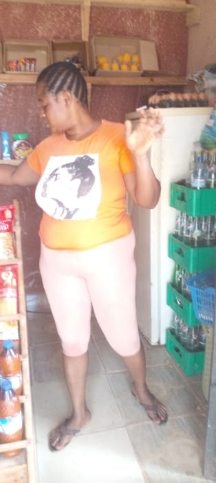 An adult female is depicted standing inside a retail shop located in Agunmo, Nigeria. She is positioned centrally, facing slightly left, and wears a short-sleeved orange t-shirt with a white graphic print, light pink capri-length leggings, and dark-colored flip-flops. Her hair is styled in cornrows, and her right arm is bent at the elbow with her hand raised, fingers slightly spread. To her left, a wooden shelving unit contains various packaged goods, including bottles filled with a reddish-brown liquid. To her right, a white refrigerator is visible adjacent to a vertical stack of green plastic crates holding clear plastic bottles. Above the refrigerator, a wooden shelf displays a carton of eggs and a cardboard box containing multiple small yellow packages. The background wall features a reddish-brown textured finish, and the floor is covered with light-colored square tiles.
