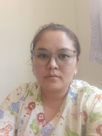 A portrait-style, head-and-shoulders photograph shows a woman wearing glasses and a light-colored shirt with a cartoon animal print. The woman has dark hair pulled back from her face. She is wearing large, rectangular glasses with clear frames. Her shirt has a pattern of various animals, including lions, frogs, elephants, mice, sheep, and cats. The background is a plain, light-colored wall, with a hint of patterned fabric, possibly a curtain, on the right side. The lighting is soft and even. The overall impression is of a casual, indoor setting. The location is the city of Kaarman, Kyrgyzstan.