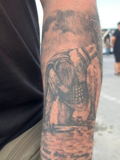 A close-up, slightly angled, outdoor shot shows a tattooed forearm of a person wearing a black t-shirt and khaki pants. The tattoo, rendered in black ink, depicts a figure hunched over a structure. The figure appears to be wearing a helmet and some form of armor or layered clothing. The lower portion of the tattoo shows a body of water with what looks like ripples or waves. Faint, blurry figures can be seen in the background, suggesting an urban environment. The person's forearm is hairy. The location is identified as Palanca, Moldova.