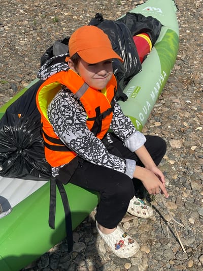 A young person wearing an orange cap and an orange life vest is sitting on a green inflatable kayak on a gravelly riverbank in Samorodovo, Russia. The kayak is labeled "AQUA MARINA". Black trash bags are piled on the kayak. The person is wearing black pants and white clogs decorated with Mario characters. The person's hands are resting on their lap. The background is a mix of rocks and sparse vegetation.