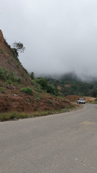 The image depicts a scenic view of a winding road passing through a mountainous, forested area with a light mist or fog. On the left, a steep, eroded slope covered with brown soil and sparse vegetation rises above the road. The road itself is paved and appears to be in good condition. In the middle ground, a white SUV is parked on the side of the road. Beyond the road, a lush green forest stretches across the hills, partially obscured by a soft, grey mist that also blankets the distant mountain peaks. Utility poles with wires are visible on the right side of the image, suggesting proximity to some form of infrastructure. The overall atmosphere is serene and natural, with a sense of elevation and expansive views.