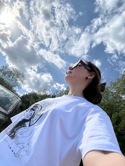 A low-angle outdoor shot depicts a woman in a white graphic tee and sunglasses. She is looking down, with a glimpse of her ear and dark hair pulled back. The background features a blue sky dotted with white clouds, interspersed with green treetops. A portion of a black car is visible at the bottom. The location is most likely not in the city of Doksha, Russia, as there is nothing in the image to indicate that.