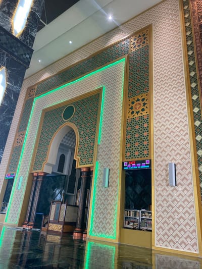 The image shows a section of the interior of a mosque, characterized by ornate Islamic geometric patterns and gold accents. The wall is decorated with a repeating pattern of beige diamond shapes with small red squares in their centers, framed by geometric borders in green and gold. A large, arched doorway is central to the image, adorned with intricate green and gold patterns. Green LED lights trace the edges of the doorway and the surrounding architectural features, creating a vibrant glow.

To the left of the doorway, a large marble-clad column with dark veining is visible, extending upwards towards the ceiling which features bright, rectangular light fixtures. To the right of the doorway, a digital display shows prayer times and dates. Below this display, a shelf unit filled with books is visible.

In the foreground, the highly polished floor reflects the architectural details and the green lighting. A wooden minbar, a raised pulpit, is positioned within the alcove of the doorway. Dark, decorative columns with copper-colored capitals support the upper structure. The overall impression is one of grandeur and meticulous craftsmanship. The location is identified as Aluekambuk, Indonesia.
