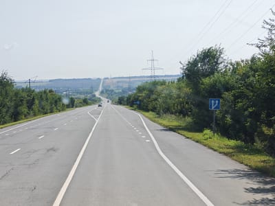 The image shows a highway in Berezki, Russia, on a bright, hazy day. The road is asphalt with white lane markings, some of which are faded or cracked. There are two lanes of traffic in each direction. Trees and green foliage line the sides of the road, and in the distance, power lines and pylons are visible, stretching across rolling hills and agricultural fields. A blue traffic sign with two upward-pointing arrows is posted on the right side of the road. A few cars are present on the highway, appearing to be in motion. The overall scene suggests a rural or semi-rural highway environment.
