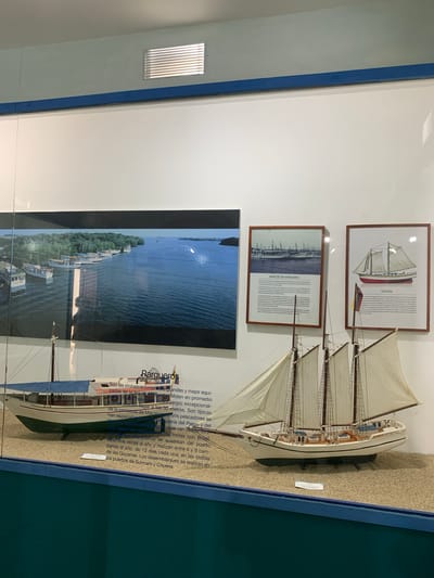 The image displays a museum exhibit featuring model ships and framed informational posters. In the foreground, two model boats are positioned on a sand-covered surface, representing a diorama. The boat on the left is a larger, motorized vessel with a blue canopy, while the boat on the right is a three-masted sailing schooner. Behind the models, a large photograph depicts a body of water lined with several boats and a forested coastline. Two framed posters are mounted on the wall to the right of the photograph. The left poster is titled "BARCOS DE MARGARITA," and the right poster is titled "GOLETA." The exhibit appears to be in an indoor setting with a white wall and a blue accent stripe running along the top. The overall lighting suggests an indoor display environment. The scene is located within the Museo de Navegación in Boca del Río, Venezuela.