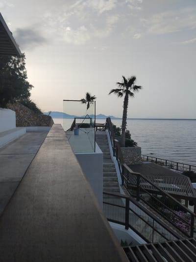 The image depicts a view of the sea and sky from an elevated location in what seems to be a coastal area. The sky is pale with light cloud cover. The sea is calm and stretches to the horizon.

In the foreground, there's architectural detail. Steps and walkways made of light-colored concrete are visible. A modern railing made of metal and glass is present. A palm tree is seen through the glass railing.

The picture appears to be taken in a location that overlooks the sea, possibly a resort or residential property with an outdoor living area or balcony. The presence of palm trees suggests a warm climate. The city of Pyli, Greece is the location.