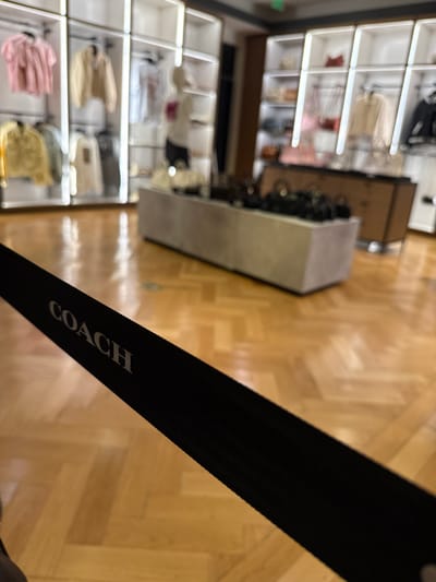 The photo depicts an overhead shot of a Coach retail store, situated in Sunrise, Florida, United States. Display shelves laden with merchandise, including bags and other accessories, are visible. A black strap with the word "COACH" printed on it is prominently in the foreground. The flooring appears to be light brown, possibly wooden, and provides a clear contrast to the black strap.